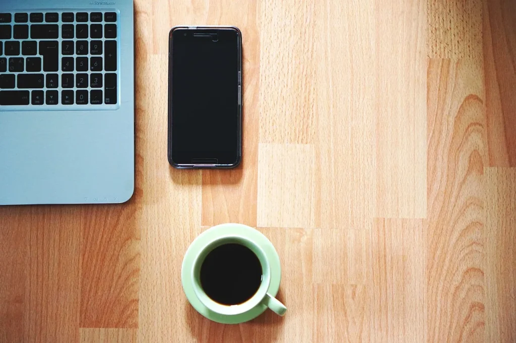 An overhead shot of a light wooden desk featuring the top-left corner of a laptop, a black smartphone, and a green cup of coffee with its saucer, symbolizing a modern, interconnected workspace.