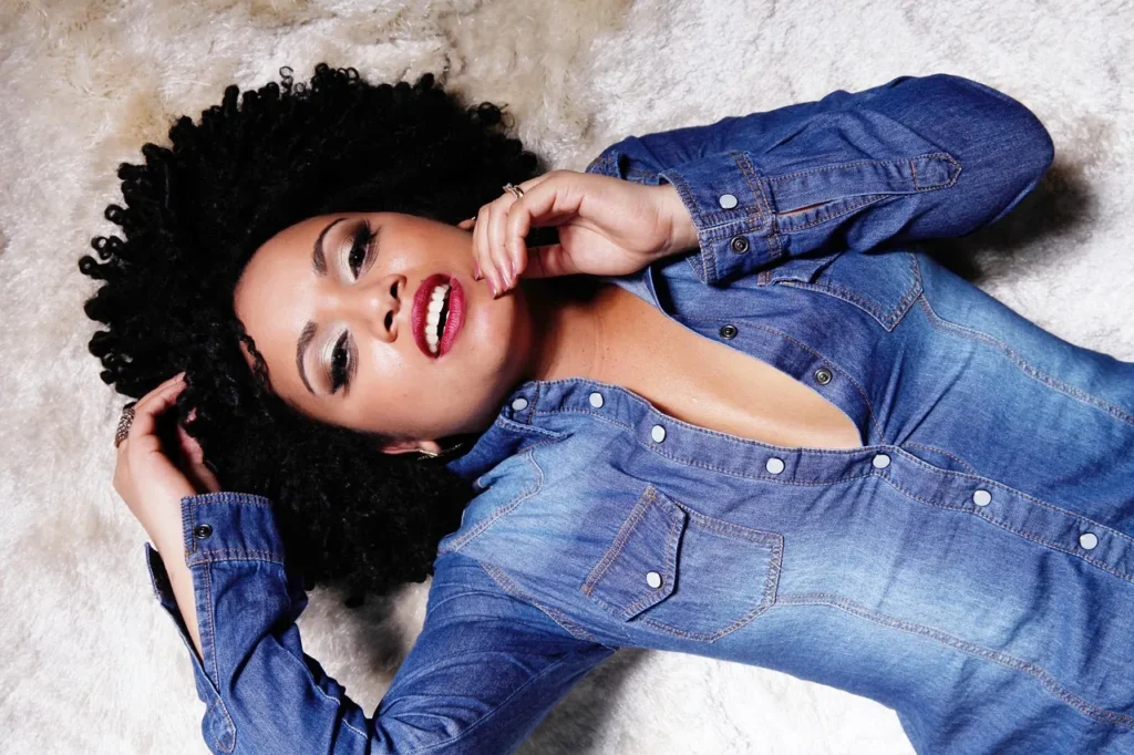 A striking, high-angle portrait of a woman with voluminous, tightly coiled natural dark hair, wearing a denim shirt, and looking confidently up at the camera.