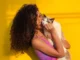 A striking portrait of a woman with long, voluminous, dark brown curly hair and a pink top, holding and gently kissing a white and black Ragdoll cat against a vibrant yellow background.