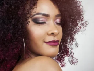 A close-up, high-fashion portrait of a woman with vibrant, deep plum-colored, tightly coiled curly hair and dramatic eye makeup, looking over her shoulder with her eyes gently closed.