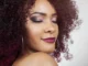 A close-up, high-fashion portrait of a woman with vibrant, deep plum-colored, tightly coiled curly hair and dramatic eye makeup, looking over her shoulder with her eyes gently closed.