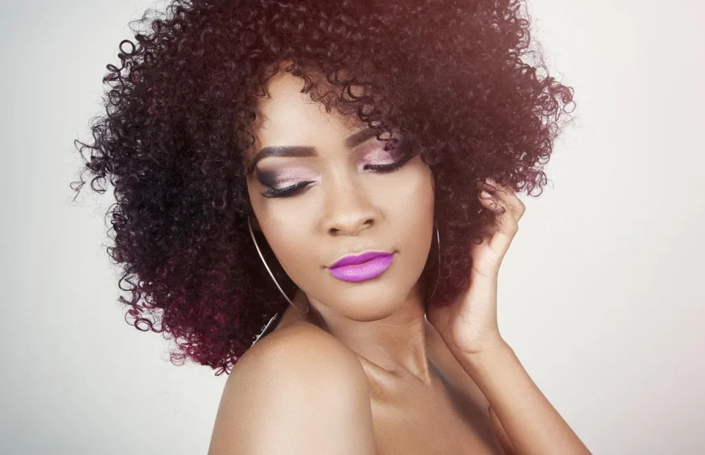 A close-up portrait of a woman with vibrant purple, tightly curled hair and metallic pink eye makeup, gently holding a lock of hair and looking down.