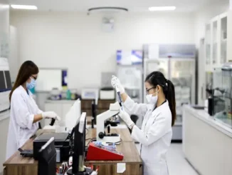 Two female scientists or lab technicians wearing white lab coats, face masks, and gloves are diligently working with laboratory equipment, including a microscope and test tube rack, in a clean, modern medical lab setting.