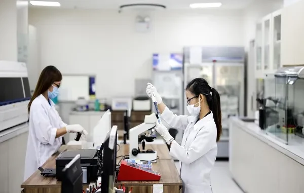 Precision and Trust: The Data Foundation of Modern IVF Two female scientists or lab technicians wearing white lab coats, face masks, and gloves are diligently working with laboratory equipment, including a microscope and test tube rack, in a clean, modern medical lab setting.