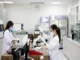 Two female scientists or lab technicians wearing white lab coats, face masks, and gloves are diligently working with laboratory equipment, including a microscope and test tube rack, in a clean, modern medical lab setting.