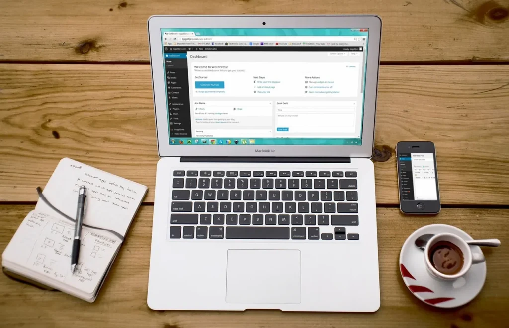 An overhead view of a wooden desk with a MacBook Air displaying a website dashboard, an iPhone, a notebook, and a cup of coffee, representing a busy, connected digital workspace.