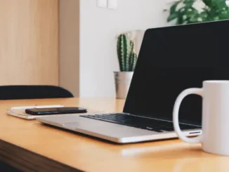 A side-angle shot of a minimalist wooden desk with an open laptop, a smartphone, and a white mug, symbolizing a modern, interconnected workspace.