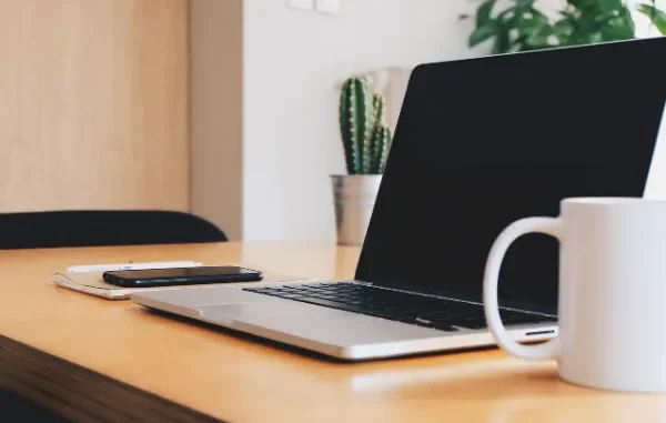 Achieving Seamless Device Connection: Your Phone Link Troubleshooting Guide A side-angle shot of a minimalist wooden desk with an open laptop, a smartphone, and a white mug, symbolizing a modern, interconnected workspace.
