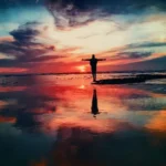 A silhouette of a person standing at the edge of the ocean with arms outstretched against a dramatic, colorful sunset or sunrise sky reflecting brightly on the wet sand and water, symbolizing freedom and recovery.