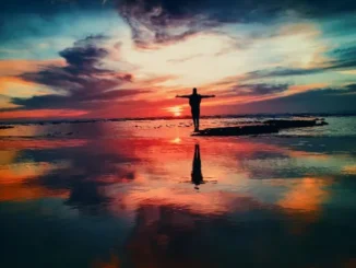 A silhouette of a person standing at the edge of the ocean with arms outstretched against a dramatic, colorful sunset or sunrise sky reflecting brightly on the wet sand and water, symbolizing freedom and recovery.
