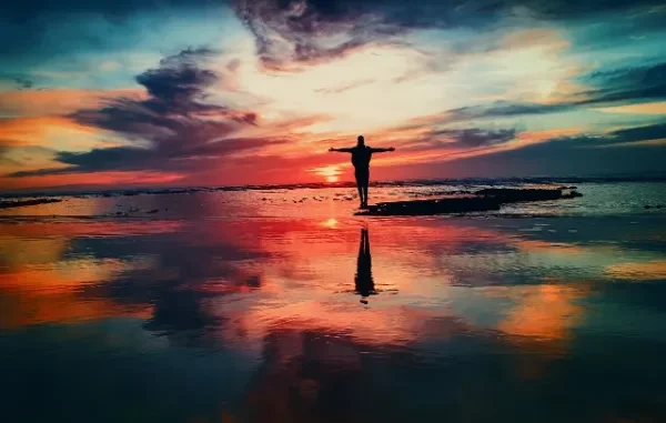 Finding Your Horizon: Regaining Control on the Road to Recovery A silhouette of a person standing at the edge of the ocean with arms outstretched against a dramatic, colorful sunset or sunrise sky reflecting brightly on the wet sand and water, symbolizing freedom and recovery.