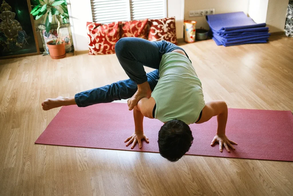 A person in a mint green tank top is performing an advanced yoga arm balance pose, possibly the Peacock or Bakasana variation, on a red mat in a brightly lit studio.