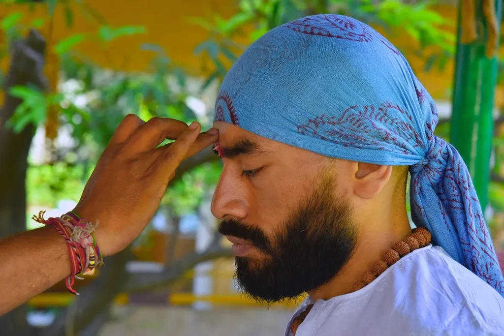 A close-up of a bearded man with his eyes closed and a blue head covering, as another person applies a red mark (tilak or bindi) to his forehead, symbolizing spiritual devotion and focus.