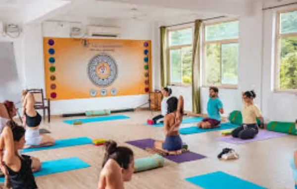 Inner Focus: A Yoga Class in Rishikesh Students are seen practicing a seated yoga pose, the Gomukhasana (Cow Face Pose) in a bright, modern yoga studio with large windows and an orange banner displaying circular chakra graphics on the back wall.