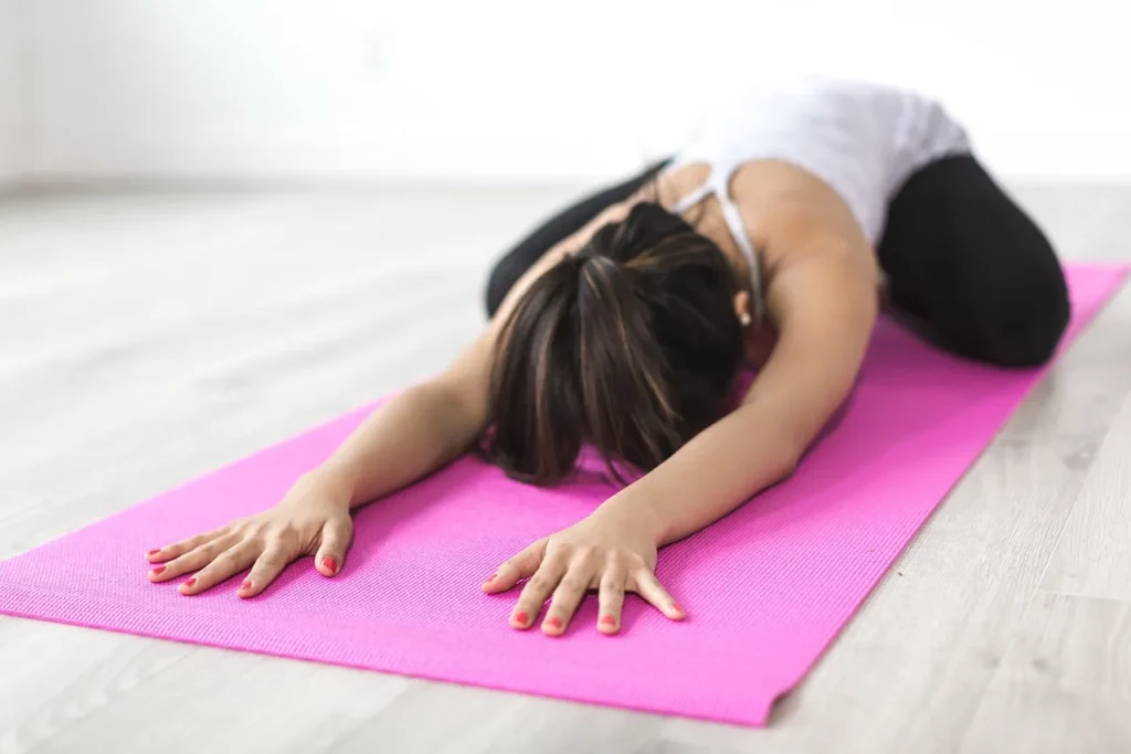 A woman in a white tank top and black pants is resting in the Balasana (Child's Pose) on a bright pink yoga mat on a light wooden floor, demonstrating focus and restorative practice.