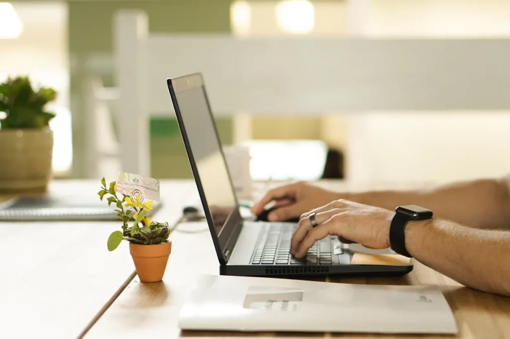 A person's hands typing on a black laptop keyboard, with a small potted plant and an envelope on a light wooden desk, symbolizing work and a touch of nature.