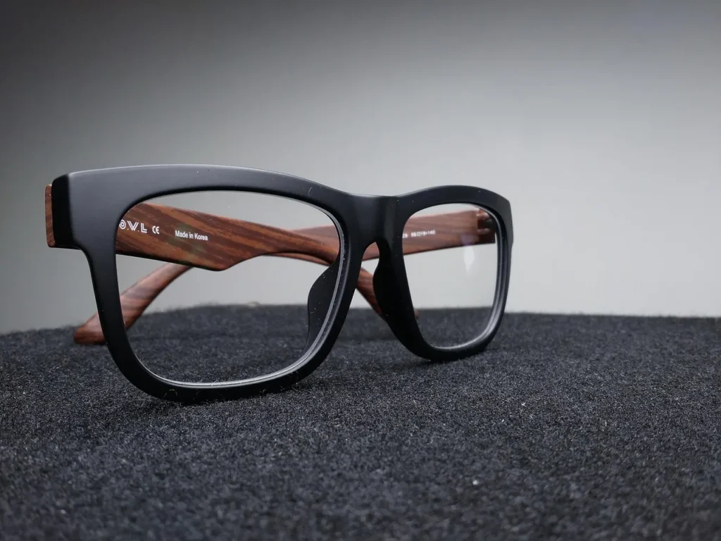 A close-up, low-angle shot of a pair of modern, thick black plastic eyeglass frames with wooden arms, resting on a dark, textured surface, showcasing a bold eyewear style.