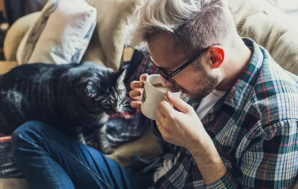 The Style Statement: Comparing Thick-Frame and Half-Frame Glasses A close-up of a man with light, styled hair and black, square-framed glasses, sitting on a couch and gently taking a sip from a mug while a striped cat looks on.