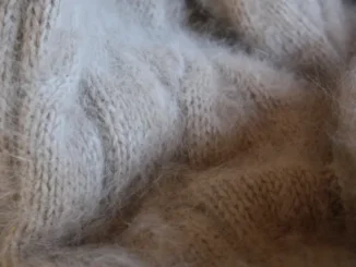 A very close-up, textured photograph of soft, light beige or tan angora or mohair knit fabric, showcasing the fuzzy, woven threads and the comforting texture of wool.