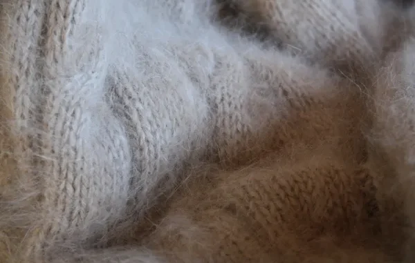 A very close-up, textured photograph of soft, light beige or tan angora or mohair knit fabric, showcasing the fuzzy, woven threads and the comforting texture of wool.