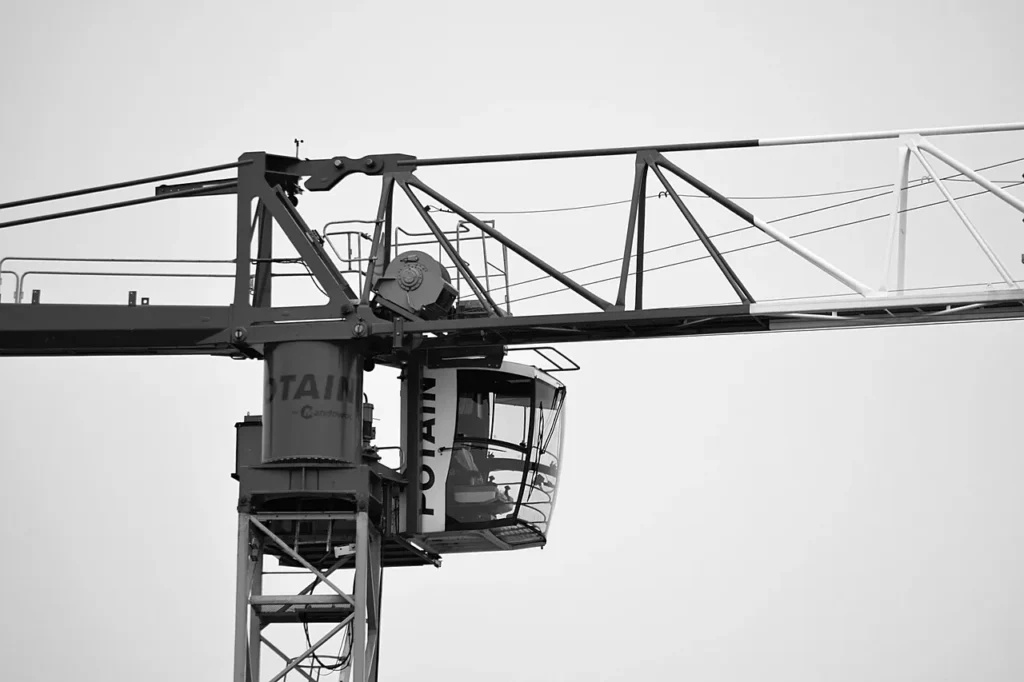 A close-up, black and white photograph of the cabin and structural boom of a large tower crane against a bright, featureless sky, representing work at significant heights.