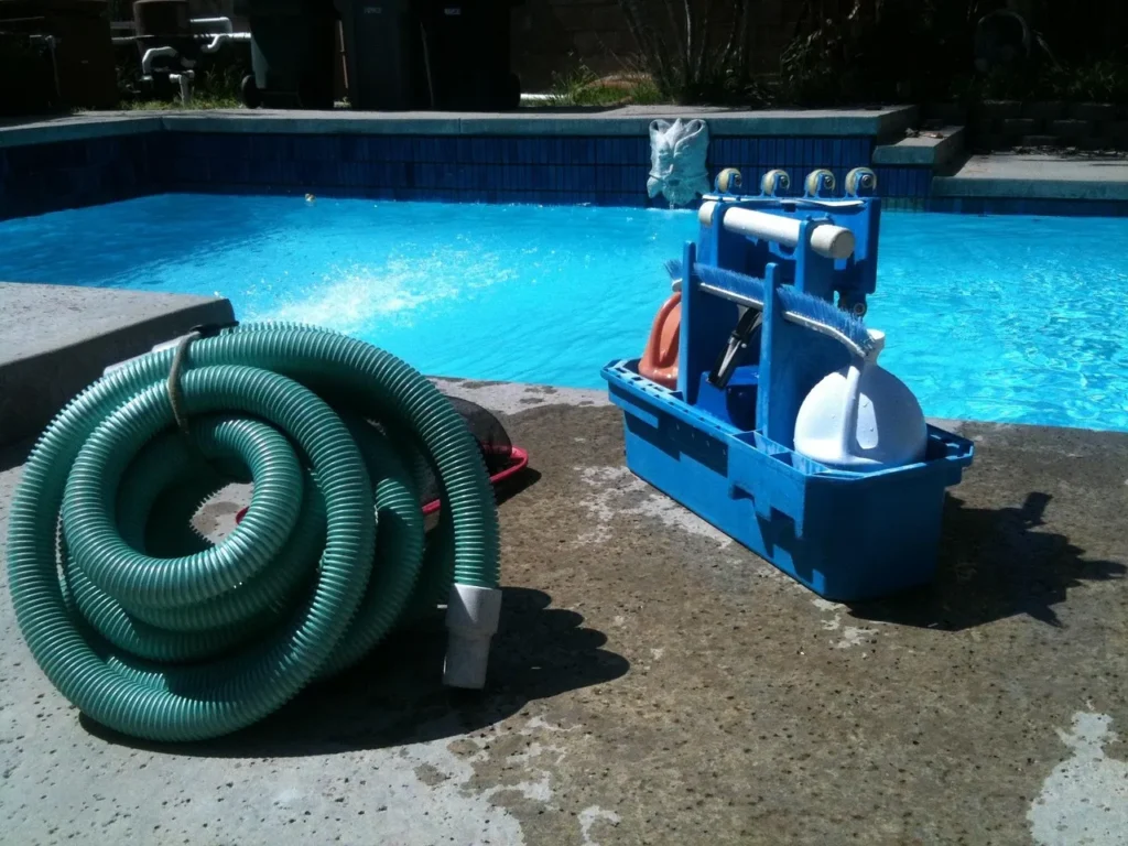 A coiled green pool vacuum hose and a blue caddy filled with traditional pool cleaning tools, including brushes and a skimmer net, sitting on the concrete edge of a bright blue swimming pool.