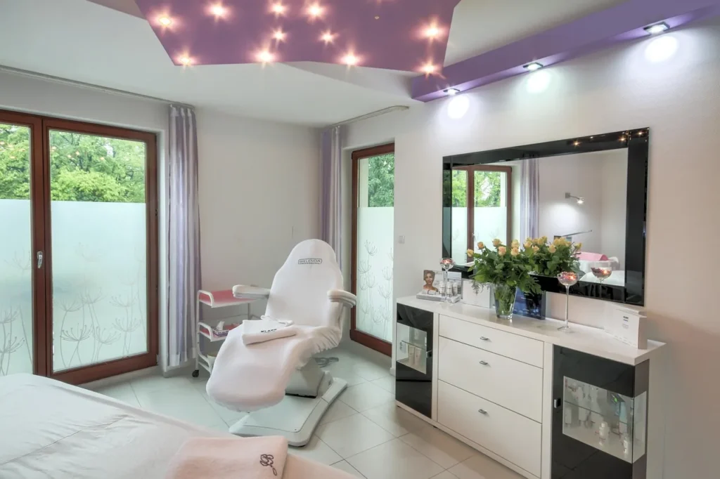 A brightly lit, modern spa or aesthetic treatment room with a white reclining chair, large mirror, elegant cabinets, and windows with frosted privacy film.