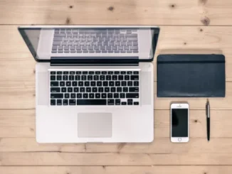 An overhead shot of a light wooden desk featuring an open laptop, a black notebook, a white smartphone, and a pen, representing a modern computing workspace.