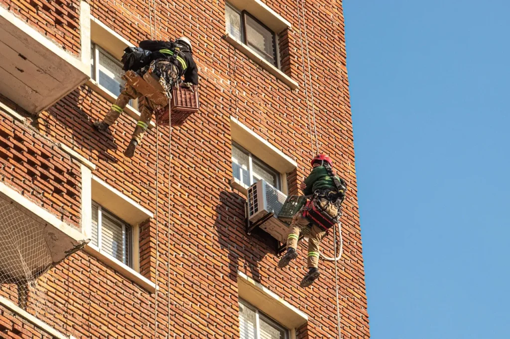 Two construction workers or technicians are suspended by ropes from the side of a tall, red brick building against a bright blue sky, performing maintenance tasks at a considerable height.