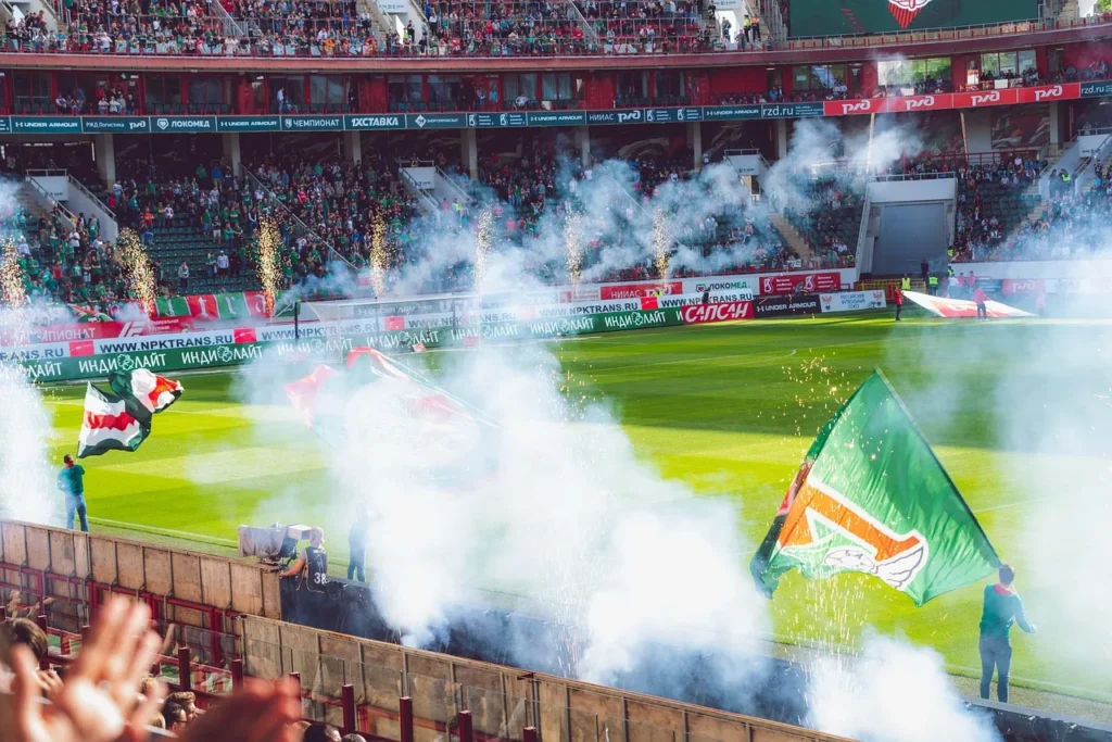 A packed football stadium during a match, featuring fans cheering, flags being waved, and pyrotechnics releasing white smoke and sparks onto the field, capturing the intense atmosphere of live football.