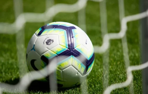 A Premier League branded football with a modern white, blue, and lime green geometric design rests on green grass behind a blurred white net, symbolizing professional soccer/football matches.