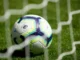 A Premier League branded football with a modern white, blue, and lime green geometric design rests on green grass behind a blurred white net, symbolizing professional soccer/football matches.