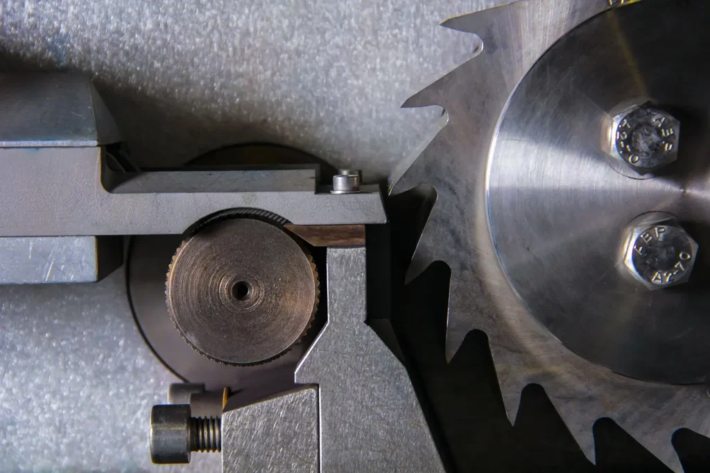 An extreme close-up of heavy industrial machinery showing a large, sharp circular saw blade positioned next to a smaller, geared metal wheel and caliper, representing the precision manufacturing of metal components.