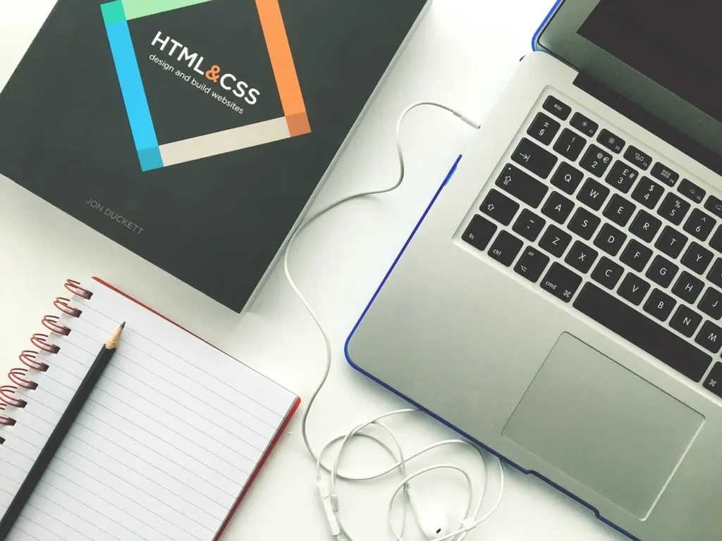 An overhead view of a clean white desk with an open laptop, a spiral notebook and pencil, and a black textbook titled "HTML & CSS design and build websites," symbolizing web development and design learning.