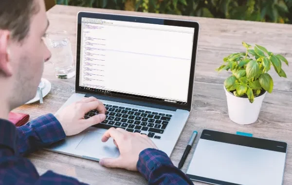 An over-the-shoulder shot of a person sitting outdoors and working on a laptop, with the screen displaying lines of code, symbolizing web development and design work using HTML templates.