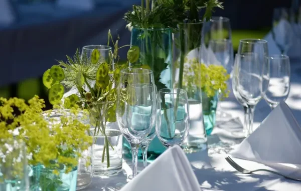A close-up of an elegant, intimate dining table setting featuring clear glassware, white folded napkins, silverware, and centerpieces of fresh green herbs and chartreuse flowers in small turquoise and clear glass vases.