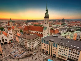 An aerial view of the historic center of Munich, Germany, at sunset, showing the Marienplatz square bustling with people and the tall central tower of the Neues Rathaus (New Town Hall) and St. Peter's Church.