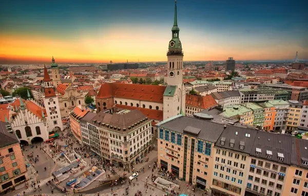 An aerial view of the historic center of Munich, Germany, at sunset, showing the Marienplatz square bustling with people and the tall central tower of the Neues Rathaus (New Town Hall) and St. Peter's Church.