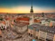 An aerial view of the historic center of Munich, Germany, at sunset, showing the Marienplatz square bustling with people and the tall central tower of the Neues Rathaus (New Town Hall) and St. Peter's Church.