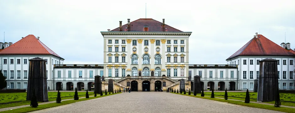 The wide, classical facade of Nymphenburg Palace in Munich, Germany, seen from the end of a long, tree-lined path, representing the historical and cultural depth of the city.