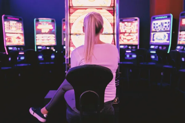 A woman with a blonde ponytail is viewed from the back, sitting intently on a chair in a dark casino setting, facing a row of brightly illuminated slot machines with pink and purple neon lights.