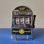 A close-up of a small, novelty toy slot machine displaying three sevens ("777") on the reels, symbolizing online slot gaming and the theme of luck and wagering.