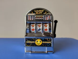 A close-up of a small, novelty toy slot machine displaying three sevens ("777") on the reels, symbolizing online slot gaming and the theme of luck and wagering.