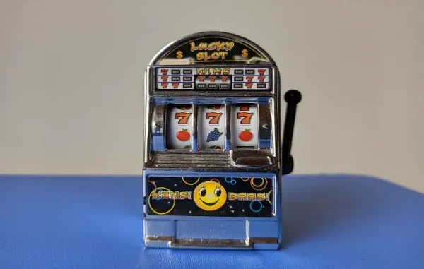 A close-up of a small, novelty toy slot machine displaying three sevens ("777") on the reels, symbolizing online slot gaming and the theme of luck and wagering.