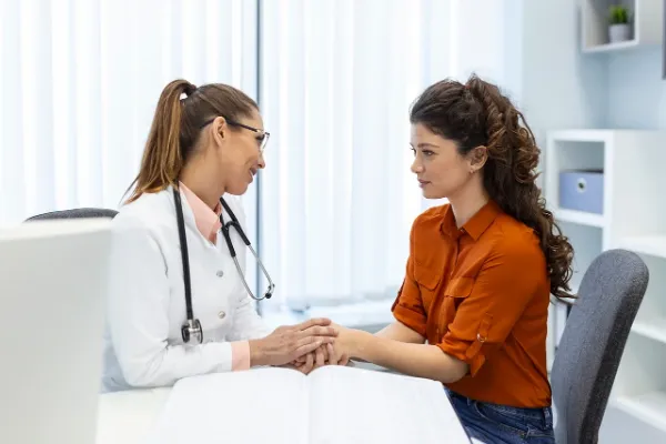 A bald, smiling male doctor wearing a white lab coat and glasses sits at a desk, gesturing with his hands while consulting with a patient (whose back is to the camera), symbolizing the medical discussion of SSRI treatment for chronic pain or PMS.