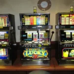 A row of three brightly lit, electronic slot machines in a casino setting, showing various credit and payout displays, symbolizing the excitement and variety of new online slot game releases.