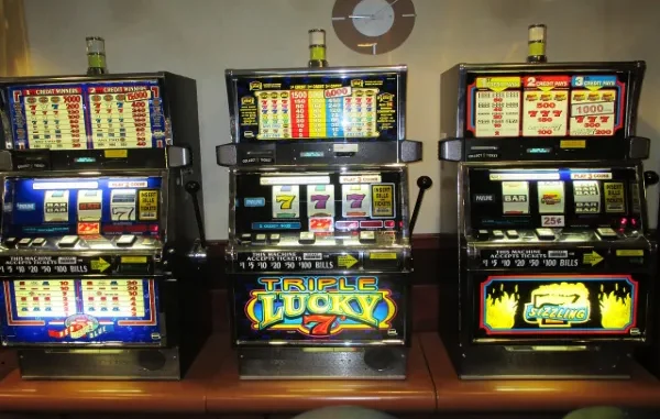 A row of three brightly lit, electronic slot machines in a casino setting, showing various credit and payout displays, symbolizing the excitement and variety of new online slot game releases.