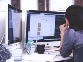A woman with dark hair sits focused at a desk with two computer monitors displaying code and web data, illustrating work in a modern office environment.
