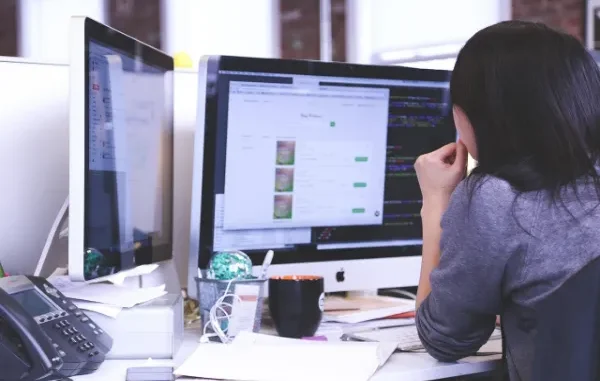 A woman with dark hair sits focused at a desk with two computer monitors displaying code and web data, illustrating work in a modern office environment.
