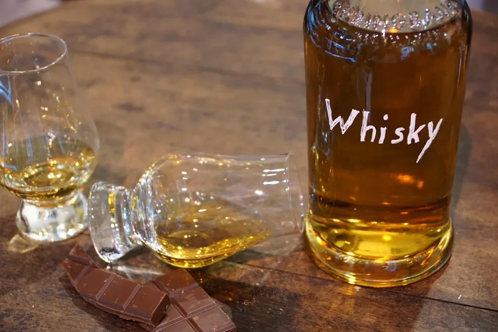 A close-up of a glass bottle of amber whiskey, labeled simply 'Whisky', alongside two tasting glasses—one lying down with liquid—and a chunk of dark chocolate on a dark wooden table, suggesting a whiskey tasting experience.